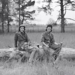 two white women in early 20th century hunting clothes perch on a fallen tree